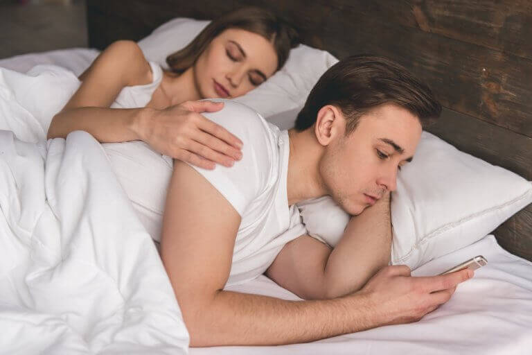 ζευγάρι στο κρεβάτι ενώ ο άνδρας ελέγχει το κινητό του, συναισθηματική απιστία